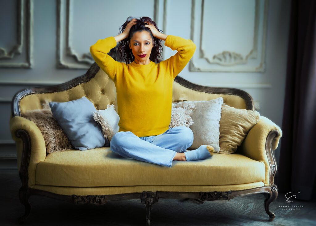 A woman with curly dark hair, wearing a yellow sweater and light blue jeans, sits cross-legged on an elegant vintage-style sofa. She poses with her hands in her hair, looking directly at the camera. The background and furniture are AI-generated, seamlessly blended with the real model.